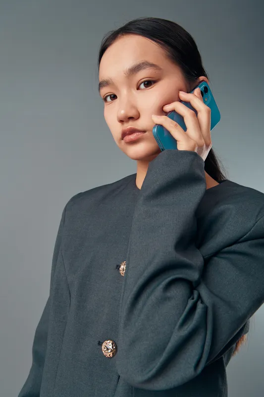 Young woman with long dark hair talking on blue smartphone, wearing oversized dark blazer with gold buttons, posing confidently in studio with plain background.
