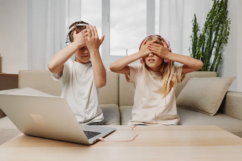 Two children with headphones play a game covering their eyes sitting on sofa with laptop on wooden table. Concept of fun, technology, and family leisure time.