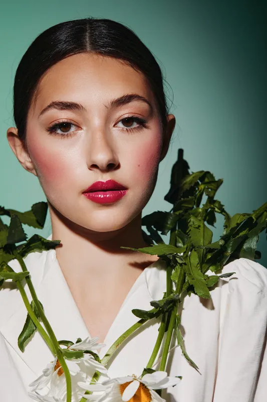 Portrait of a young woman with flawless skin and bold bright lipstick holding green plants while wearing a white outfit. Studio shot with blue background. Beauty fashion concept.