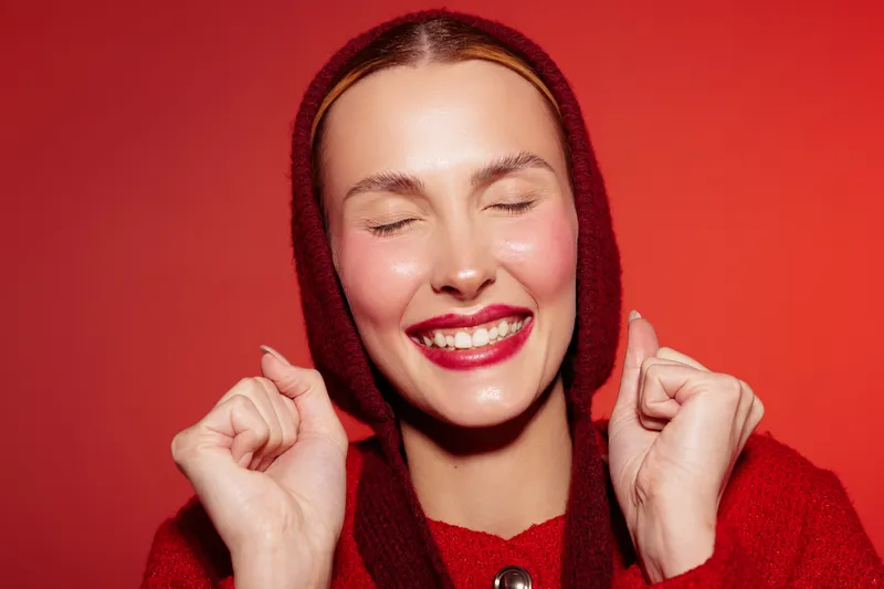 Happy woman with closed eyes smiles brightly wearing red hooded sweater on red background. Concept of joy, happiness, positive emotions, and winter fashion portrait.
