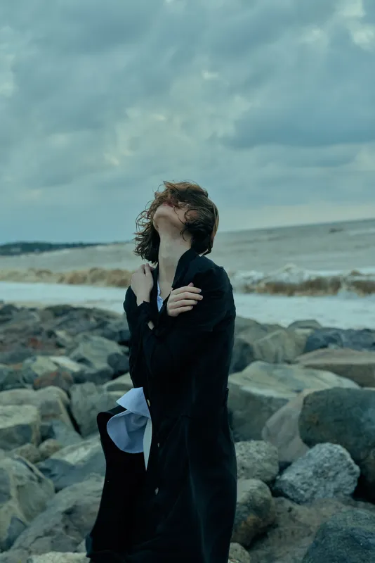 Windy seaside fashion scene with a woman in a dark coat on a rocky shore, hair swept by gusts, moody sky and crashing waves
