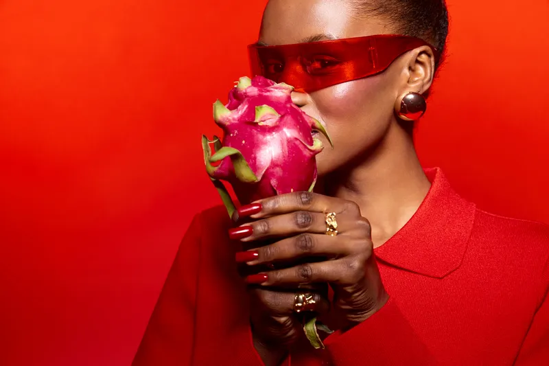 Dark skinned African woman in red clothes and red sunglasses holds dragon fruit near face on red background. Concept for fashion, beauty, fresh fruit, and vibrant color contrast.