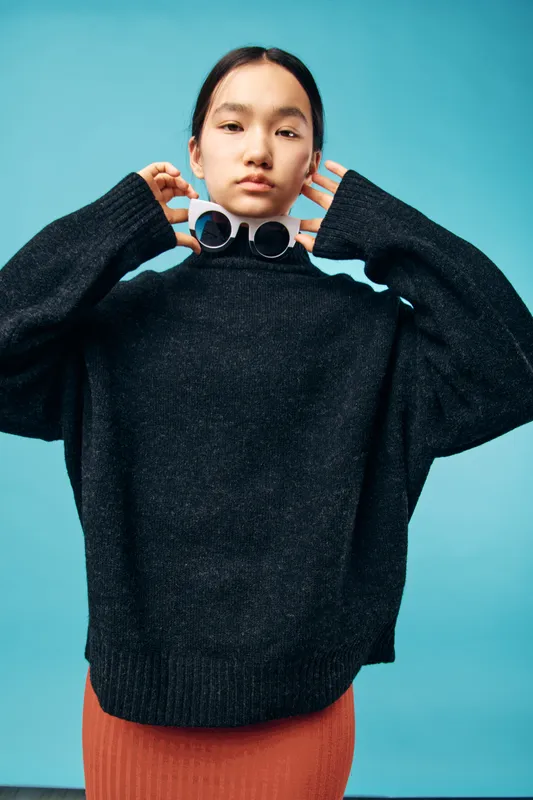Young woman posing in black sweater and red skirt against blue background with sunglasses held near neck. Fashionable casual portrait with confident expression, studio light and modern style outfit.