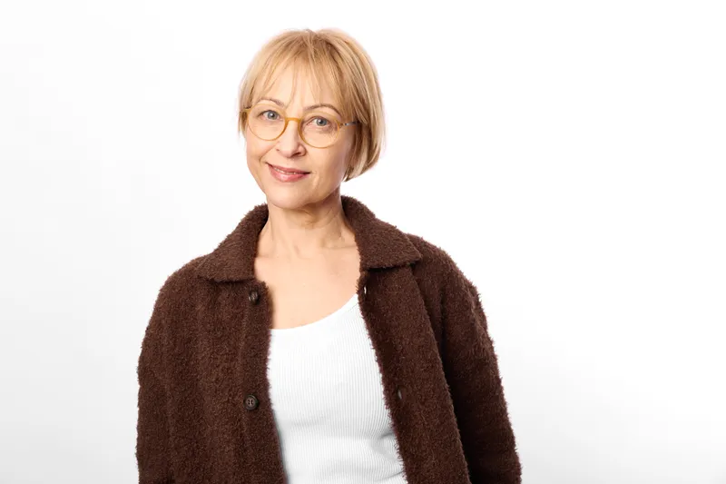 Woman with glasses, smiling portrait of an older blonde woman in a brown coat, in a studio, against a white background, warm calm mood, everyday fashion style, comfort