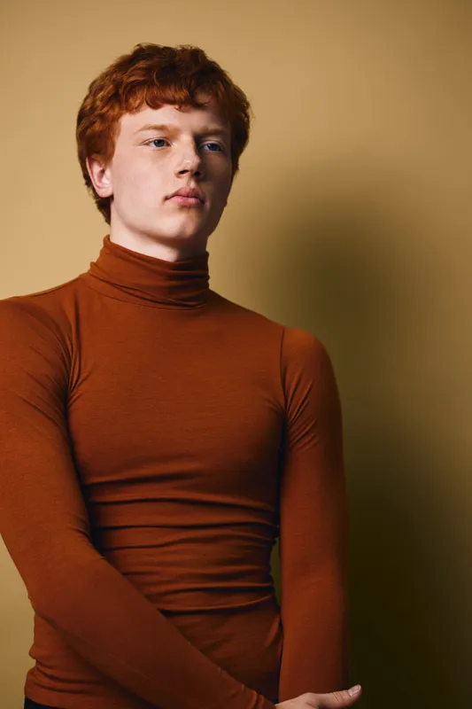 Young man with red hair wearing brown turtleneck sweater posing against plain background with serious calm expression and confident posture in studio portrait photography.