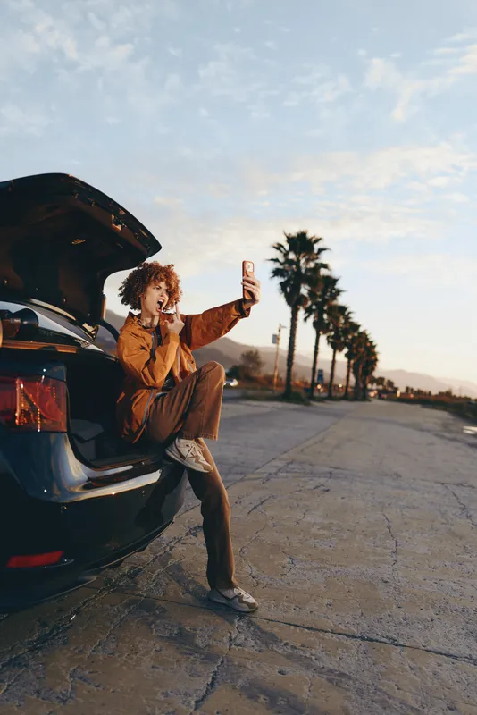 Woman lifestyle outdoors near car trunk, sitting on pavement and holding smartphone, taking a selfie at sunset, warm sky and soft light, relaxed travel mood, palm trees lining the road