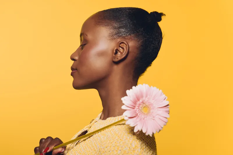 Profile of a young African woman with dark skin and natural hair tied back, holding a pink flower on a yellow background. Concept of beauty, nature, and peacefulness.