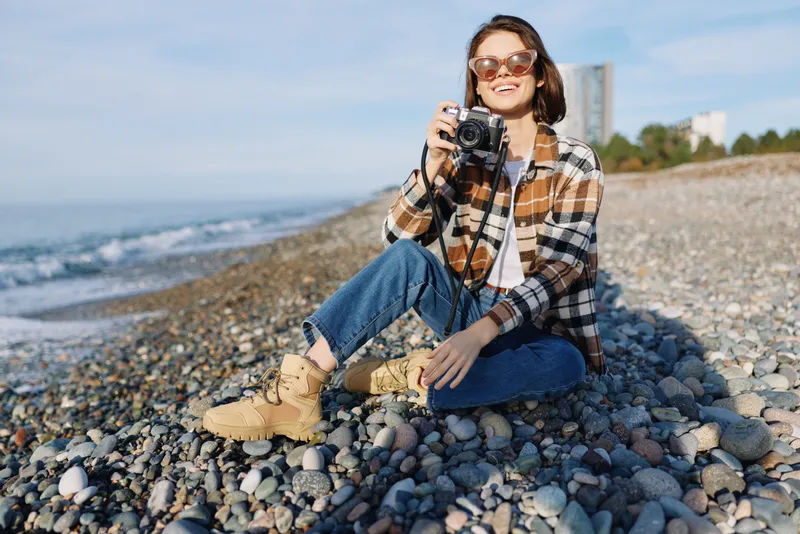 Female photographer on a pebble beach with a camera, casual plaid jacket and boots, sunny day, sea in the background, capturing moments and enjoying outdoor photography.