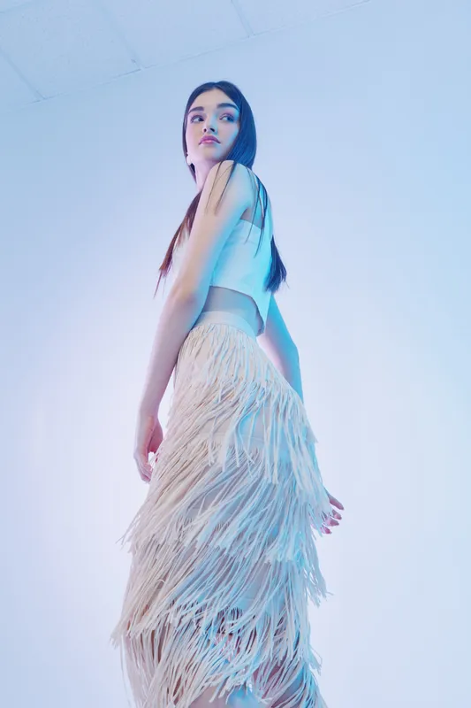 Young woman in a stylish white crop top and fringed skirt poses confidently against a light background. Concept of woman beauty fashion, elegance, and modern style.