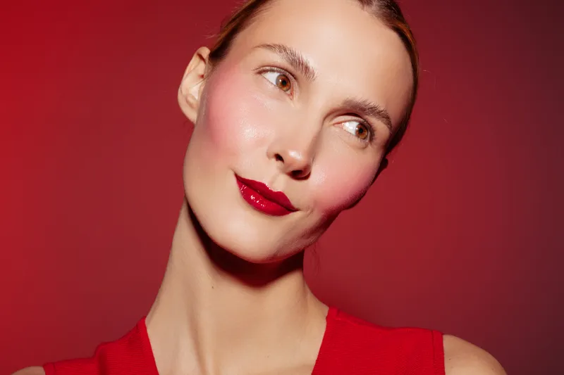 Close-up of a woman with red lipstick and smooth skin looking sideways on a red background. Concept of beauty, makeup, skincare, and fashion portrait with vibrant colors.