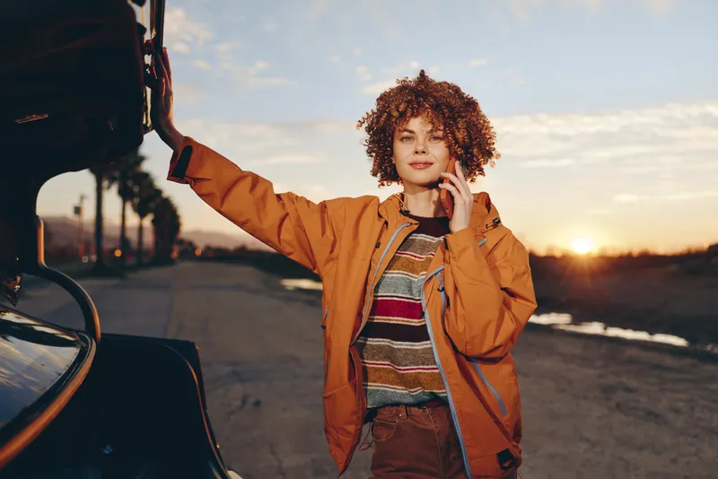Cheerful woman lifestyle outdoors by car at sunset, curly hair blowing lightly, orange jacket and striped top, relaxed confident smile, warm golden light, open road background evening