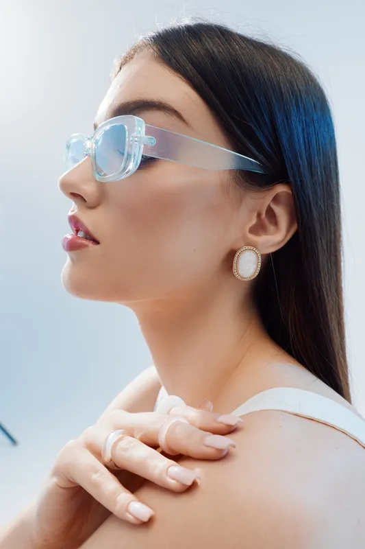 Profile of a stylish woman with long dark hair wearing transparent sunglasses and elegant earrings. The close-up highlights beauty, fashion, and modern accessories. Studio lighting.