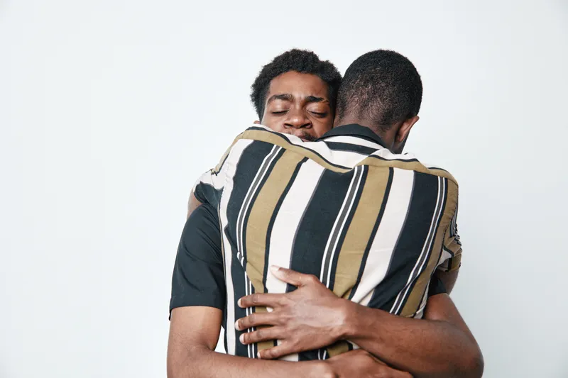 Two young black men hugging each other with eyes closed for friendship and support. Dark skin males showing affection and emotional connection on white background.