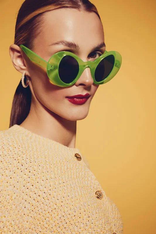 Stylish woman with green sunglasses and red lipstick poses against yellow background. She wears a textured beige sweater and gold hoop earrings. Concept of fashion, beauty, and retro style.