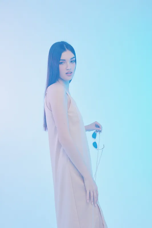 Young woman with long hair wearing a beige dress holds sunglasses in a soft blue studio light. Concept of beauty fashion elegance and modern style portrait.