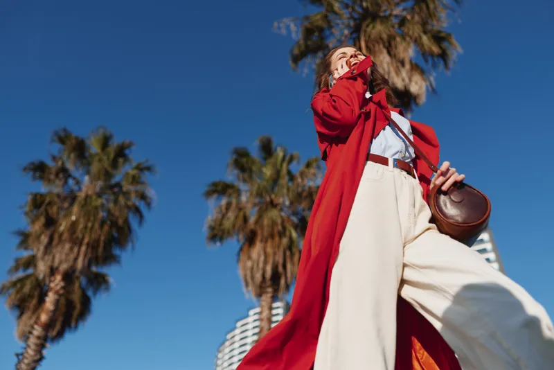 Young woman in a red coat strides outdoors under a clear blue sky, white pants swaying, a brown bag in hand, captured from a dramatic low angle.