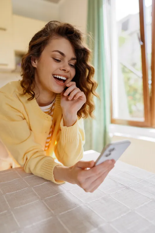 Young woman in a yellow sweater smiles at her smartphone while seated at a bright home dining table, capturing a candid moment of daily life.