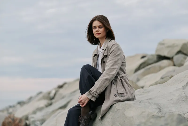 Young woman in casual coat sitting on large rocks by the sea shore. Overcast sky and calm atmosphere create a natural outdoor portrait concept of relaxation and contemplation.