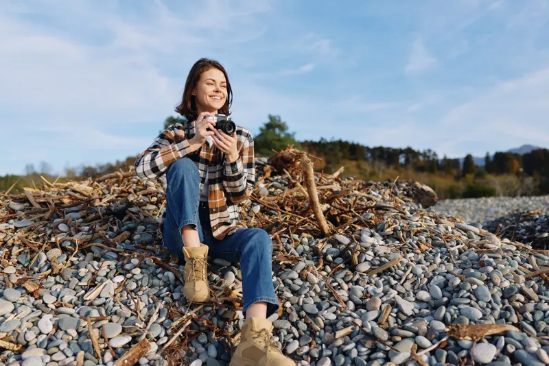 Photography, photographer, outdoors, landscape, beach, camera, nature, casual, boots, plaid woman sits on a rocky shoreline with a camera and smiles in the bright sun, driftwood and pebbles scattered