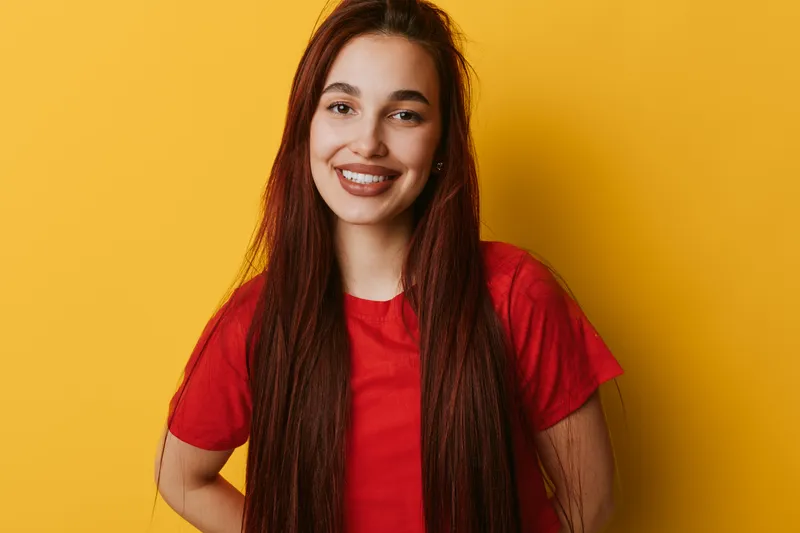 Young woman with long straight hair smiling and posing against yellow background wearing red t shirt. Portrait of happy female with natural makeup and confident expression in studio.