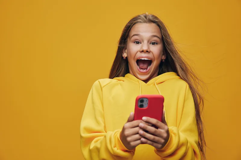 Girl with long hair wearing yellow sweater holding red smartphone showing surprised emotion with open mouth on colored background for social media or communication.