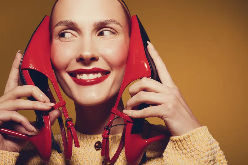 Smiling woman holds red high heel shoes near face, wearing yellow sweater. Close-up portrait on brown background. Concept of fashion, style, happiness, and beauty accessories.