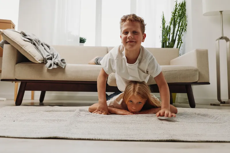 Playful siblings have fun indoors, boy smiling on girl lying on carpet in cozy living room with natural light and modern decor.