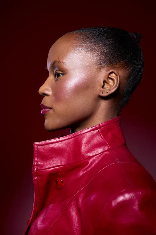 Profile of a black African woman with dark skin wearing a red leather jacket. Studio portrait with dramatic lighting on burgundy background. Concept of fashion and confidence.