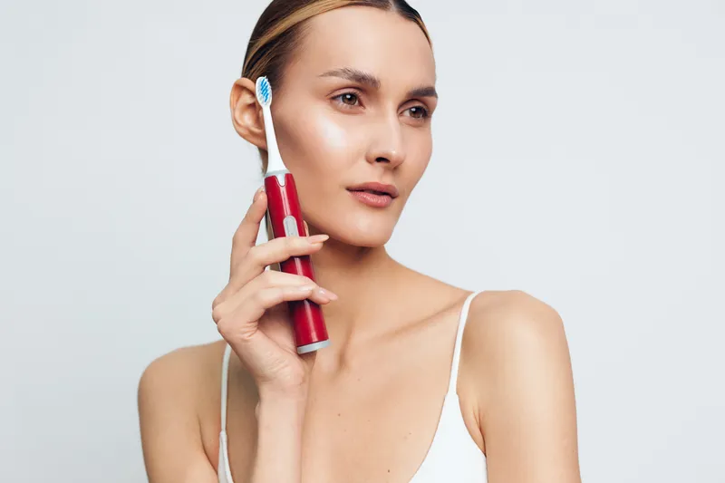 A young woman with clear skin holds a red electric toothbrush near her face against a white background. The image represents dental care, hygiene, and personal grooming.