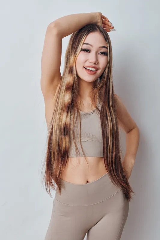 Young woman with long hair wearing beige sportswear posing with one arm raised and smiling on white background for fitness and lifestyle concept.