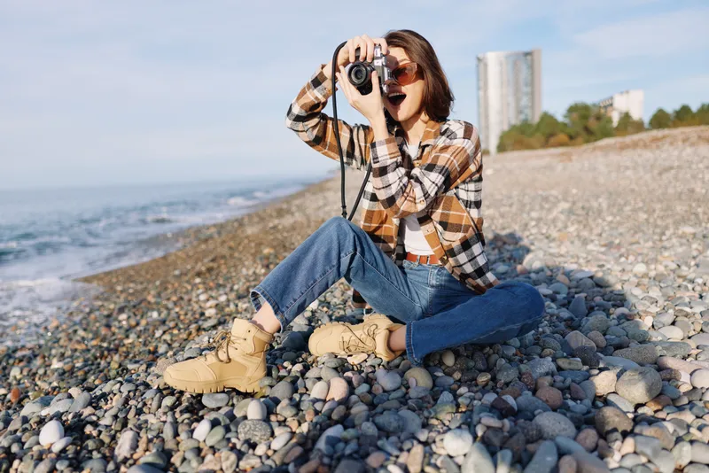 Photographer crouched on a rocky beach with a camera, scanning the shore in casual attire, capturing outdoor nature and summer exploration with focus and flair