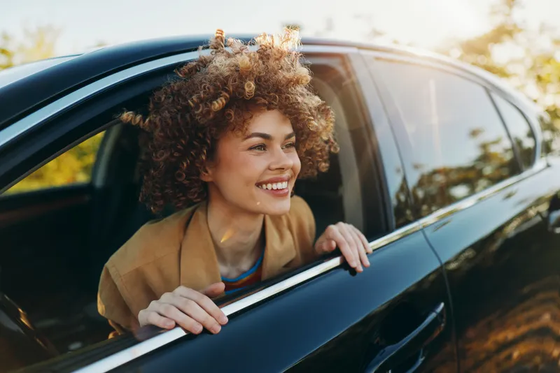 A joyful woman leans out of a car window, enjoying sunny outdoors. A candid lifestyle shot with retro film color and warm tones, capturing carefree moment and everyday happiness.