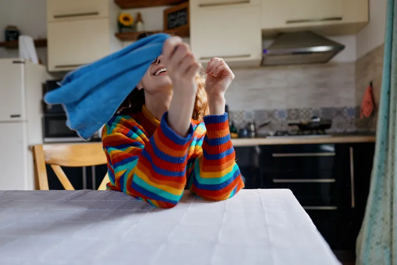 Smiling woman in a colorful striped sweater covers her face with a blue cloth at a kitchen table, capturing a playful home moment, casual domestic scene, warm mood.