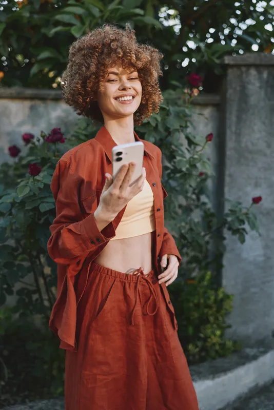 Smiling woman with curly hair holding a smartphone in an outdoor setting, rust orange outfit, sunny garden background, conveying casual fashion, lifestyle, and cheerful self expression