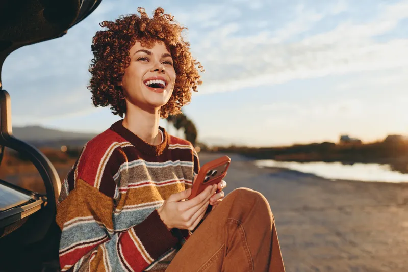 Woman lifestyle, smile with curly hair, holding smartphone near open car at beach sunset, warm evening wind, casual travel moment, candid outdoor photo
