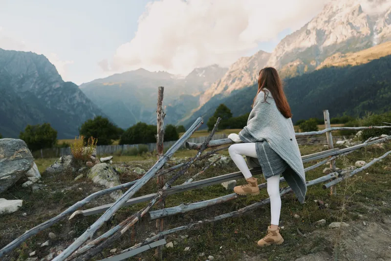 One young woman with long brunette hair wears a gray cardigan and white jeans as she leans on a rustic wooden fence in a dramatic mountain landscape.