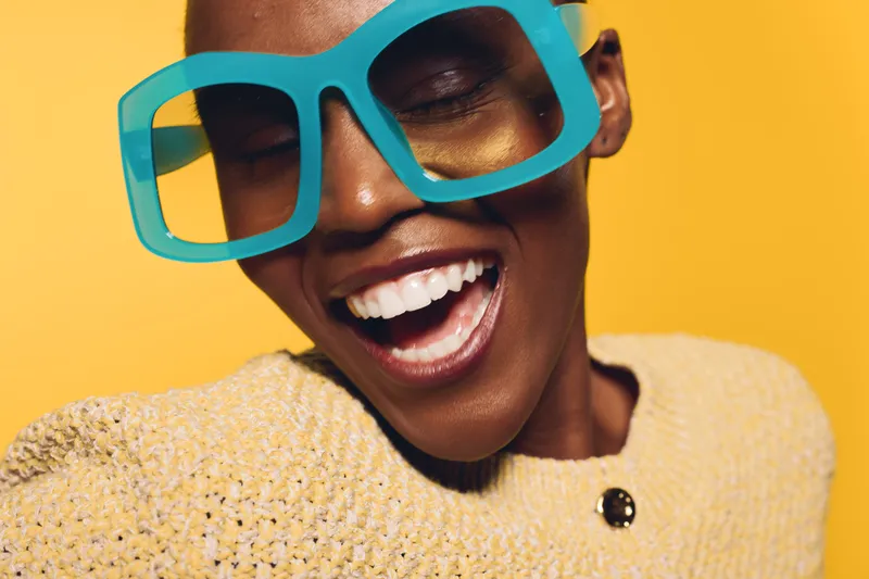 Happy African woman with dark skin wearing oversized blue glasses and yellow sweater smiles with closed eyes on bright yellow background. Concept of joy, fashion, and vibrant style.