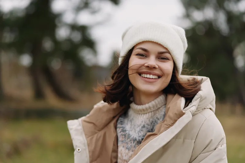 Woman outdoors in a winter coat and white beanie, smiling at the camera in a park setting, showcasing casual fashion and warm, approachable mood.