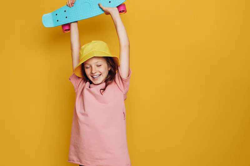 Happy child in a yellow hat raises a blue skateboard above the head. Pink shirt and bright yellow background create joyful mood. Active lifestyle concept.