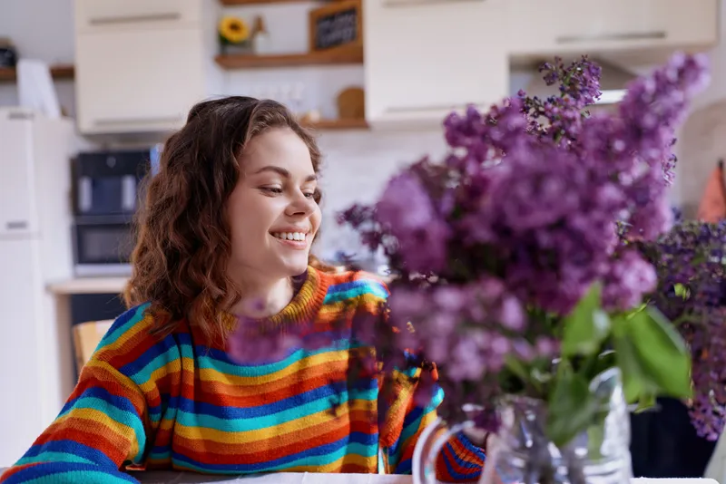 Smiling woman in a colorful striped sweater sits at home in a kitchen, surrounded by purple flowers. A candid lifestyle moment focused on everyday comfort and cozy, cheerful ambience.