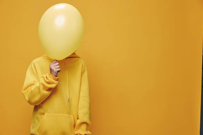 Person in yellow hoodie holding yellow balloon in front of face against yellow background for anonymity, casual style and monochrome concept in portrait.
