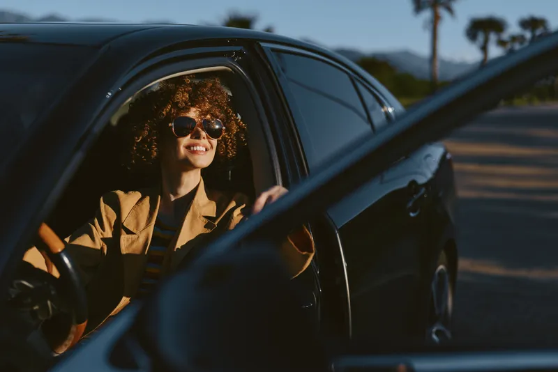 Smiling woman with curly hair sits in a car, wearing sunglasses and a blazer. Warm retro tones create a casual lifestyle mood and travel vibe.