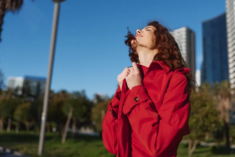 Young woman in a bright red coat stands outdoors under a clear blue sky, embracing a breeze with eyes closed and head tilted toward the sun.