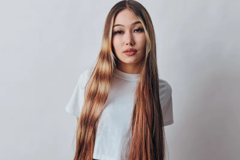 Young woman with long straight hair and casual white shirt posing in studio with neutral background, looking confidently at camera with natural makeup and relaxed expression.