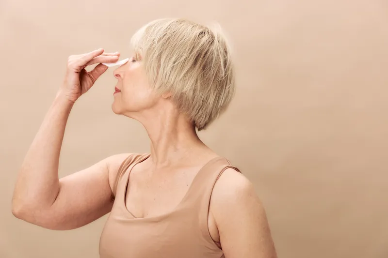 Profile of mature woman with short blonde hair pinching nose in beige tank top on neutral background. Concept of stress relief, headache, discomfort, and wellness for older adults.