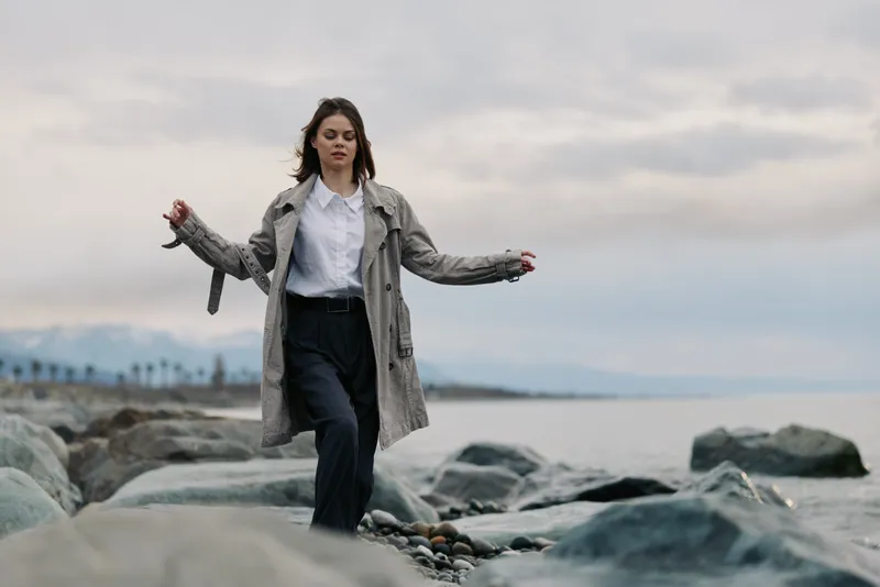 Young woman in a grey coat walks carefully on rocks by the sea under cloudy sky. Casual outfit and natural landscape create a serene and thoughtful mood.