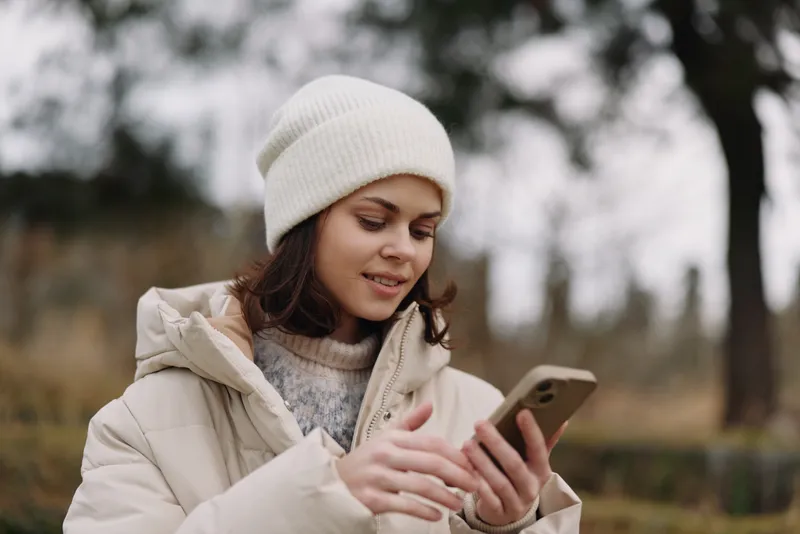 woman outdoors smartphone winter coat beanie cold, a relaxed portrait of a woman browsing content on her mobile in a park on a chilly day, capturing candid connectivity and calm urban nature