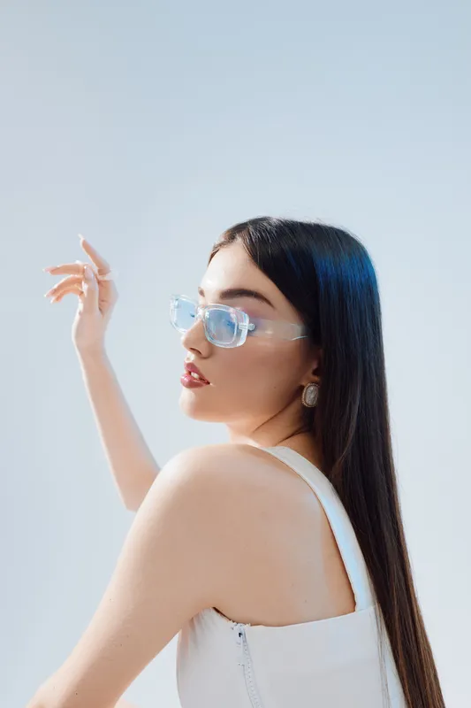 Young woman with long dark hair wearing transparent glasses and white top poses against a light background. Concept of beauty, fashion, and modern style with natural makeup.