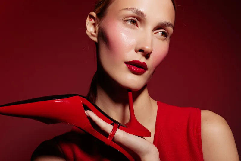 Close-up of a stylish woman with red lipstick holding a red high heel shoe near her face. Fashion concept with bold makeup and elegant accessories on red background.