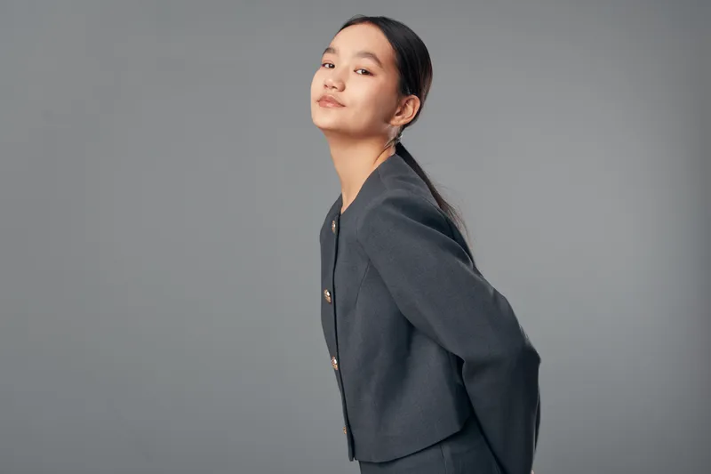 Young woman in business attire posing confidently on gray background with hands behind back. Professional female model with neat hairstyle and subtle makeup looking at camera.